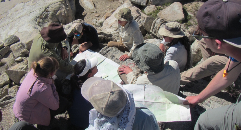 A group of outward bound students rest on rocks while examining maps. 